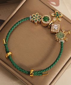 a green leather bracelet with gold charms and clovers on the clasp is sitting on a brown box