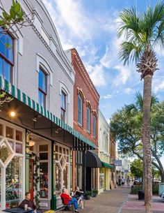 people sitting at tables in front of shops on a street with palm trees and parked cars