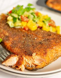 a piece of fish on a white plate with a fork and some salad in the background