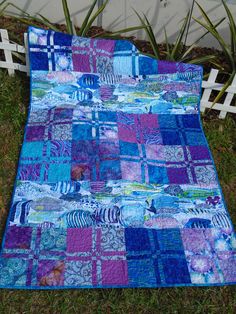 a blue and purple patchwork quilt laying on the grass next to a white fence