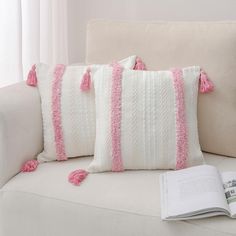 two white pillows with pink tassels and a book on a couch next to them