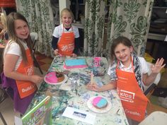 three girls in aprons standing around a table with cakes on it and holding up their hands