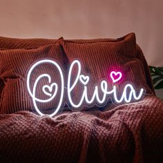 the word ollivia is lit up with neon lights on a bed in front of a potted plant