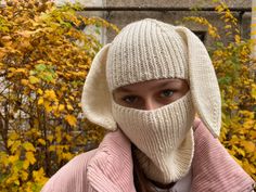 a woman wearing a knitted hat and scarf over her face in front of trees with yellow leaves