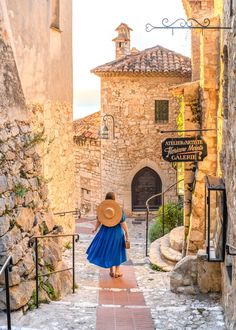 a woman in a blue dress and straw hat walks down an alleyway between two stone buildings