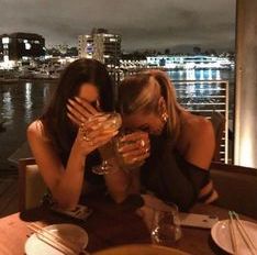 two women sitting at a table with wine glasses in their hands and city lights behind them