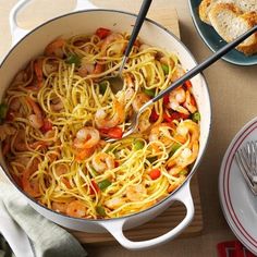 a pan filled with pasta and shrimp on top of a wooden cutting board next to bread