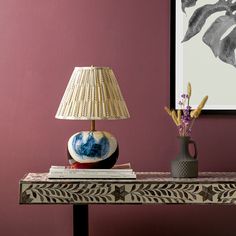 a table with a lamp, vase and books on it next to a purple wall