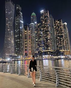 a woman is walking along the waterfront in front of some tall buildings at night time