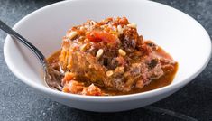 a white bowl filled with meat and sauce on top of a black table next to a fork