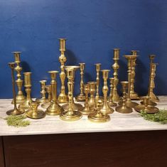 a collection of brass candlesticks and candle holders on a wooden table with blue wall in the background