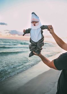 a man holding up a baby wearing a shark costume at the beach with his mouth open