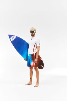 a man holding a blue surfboard while standing in front of a white background and wearing sunglasses