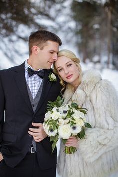 a bride and groom standing in the snow with their arms around each other, dressed in winter attire