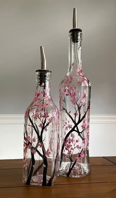 two glass bottles with pink flowers painted on them sitting on a wooden table next to each other