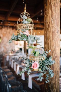 two birdcages hanging from the ceiling with flowers and greenery in them on tables