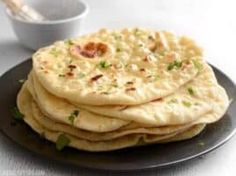 a stack of pita bread on a black plate next to a white bowl and utensils