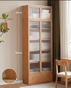 a tall wooden cabinet with glass doors next to a table and chair in front of a window