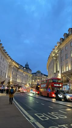 two double decker buses driving down a street next to tall buildings and traffic lights at night