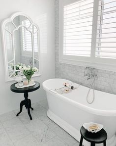 a white bath tub sitting under a window next to a table with flowers on it