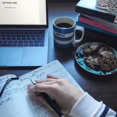 a person writing on a notebook next to a plate of cookies and a cup of coffee