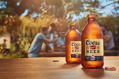 two bottles of beer sitting on top of a wooden table next to another bottle with people in the background