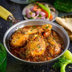 a metal bowl filled with chicken curry next to bread and veggies on a table