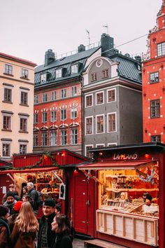 people are walking around an outdoor market with red booths and buildings in the back ground