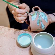 a person holding a paintbrush next to a small vase and two bowls on a table