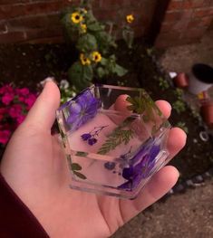 a hand holding a glass block with purple flowers in it and green leaves on the inside