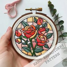 a hand embroidery pattern with roses in a stained glass frame on a white table next to some green leaves