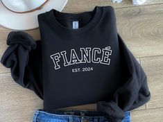 Introducing our chic and adorable Fiancé Sweatshirt - the perfect addition to your wardrobe, designed for the stylish bride-to-be! Crafted with love and attention to detail, this crewneck sweater features the word "Fiancé" in a bold varsity outlined font, available in classic black or white. To add a touch of personalization, we've included the special touch of EST 2024 in a subtle font beneath the arched "Fiancé," making this sweatshirt a timeless keepsake for your engagement year. Elevate your Fiance Clothes, Feyonce Sweatshirt, Sweatshirt Bachelorette, Bachelorette Nashville, Engaged Gifts, Fiance Sweatshirt, Engagement Party Attire, Male Sweaters, Bride Top