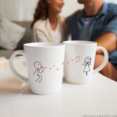 two coffee mugs sitting on top of a white table next to each other with stick figures drawn on them