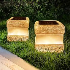 two square planters sitting in the grass next to each other with lights on them