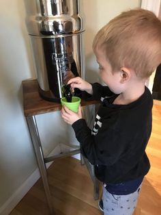 a little boy that is standing in front of a coffee pot and holding a cup