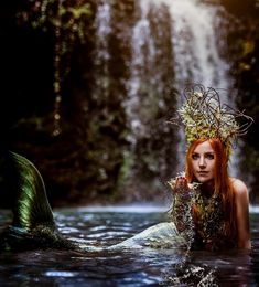 a woman with long red hair is in the water near a waterfall wearing a crown