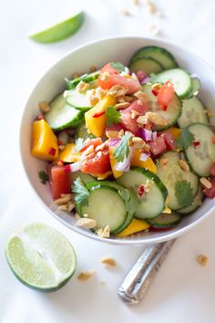 a white bowl filled with cucumber, tomatoes and other vegetables next to lime wedges