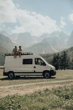 two men sitting on the roof of a white van in front of some mountains and trees