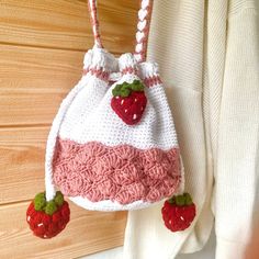 a crocheted strawberry purse hanging from a hook on a wooden wall next to a white sweater