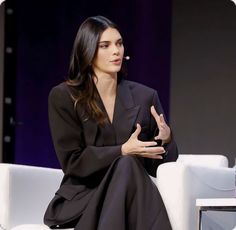 a woman sitting on a white couch talking to someone in front of her and wearing a black suit