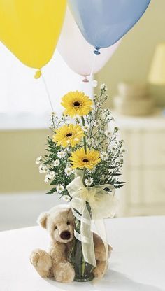 a teddy bear sitting next to a vase filled with yellow and white flowers in front of balloons