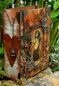 an altered book sitting on top of a rock next to plants and flowers with charms hanging from it's sides