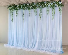 a blue backdrop with white flowers and greenery on the top, along with sheer drapes