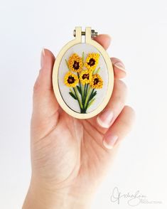 a hand is holding a small embroidery project with yellow flowers in the center and green stems