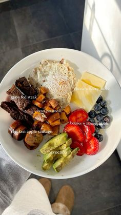 a white plate topped with meat and fruit next to a person's feet on the floor