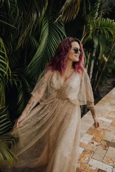 a woman with pink hair wearing a dress and sunglasses standing in front of palm trees