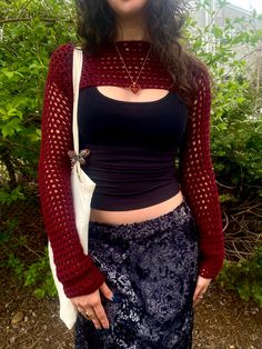 a woman with long hair wearing a black top and blue patterned skirt, standing in front of some bushes