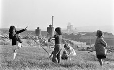 three girls are flying a kite in a field with other people watching from the hill