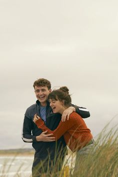 a man and woman hugging each other on the beach with tall grass in front of them