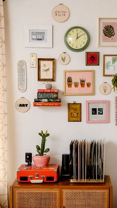 a wall with many different pictures and clocks on it's sideboard next to a plant